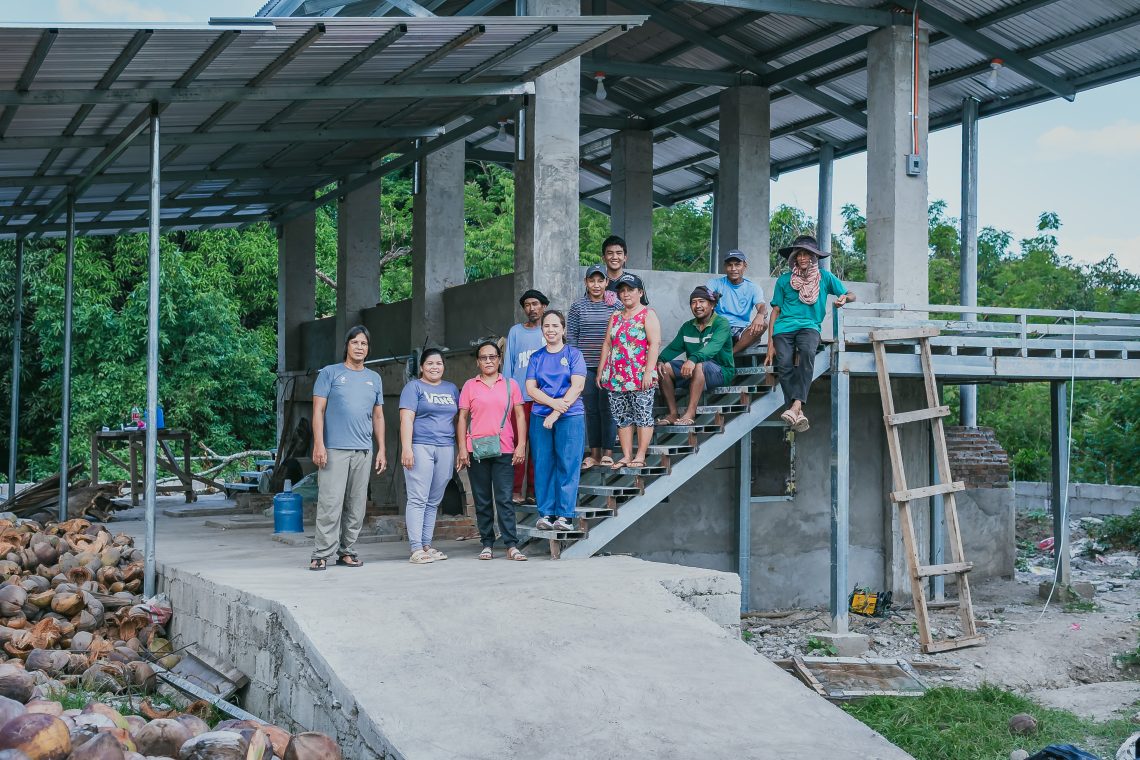 A group of men and women farmers stand on a staircase inside their coconut and copra sorting facility.
