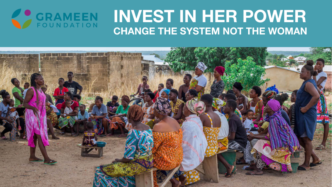 A woman stands in the center of a large group of seated people, teaching them about Grameen's power dynamics approach.  The audience is engaged with the speaker. All are outside and sitting under a tree.