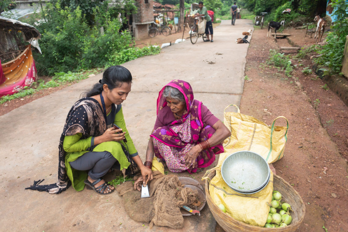 A Grameen Agent in India helps a client access digital funds using her fingerprint. The Agent is a woman in a dark blue and green sari. The client is a woman in a pink sari. They are on a dirt sidewalk, next to a basket of fruit and some burlap sacks.