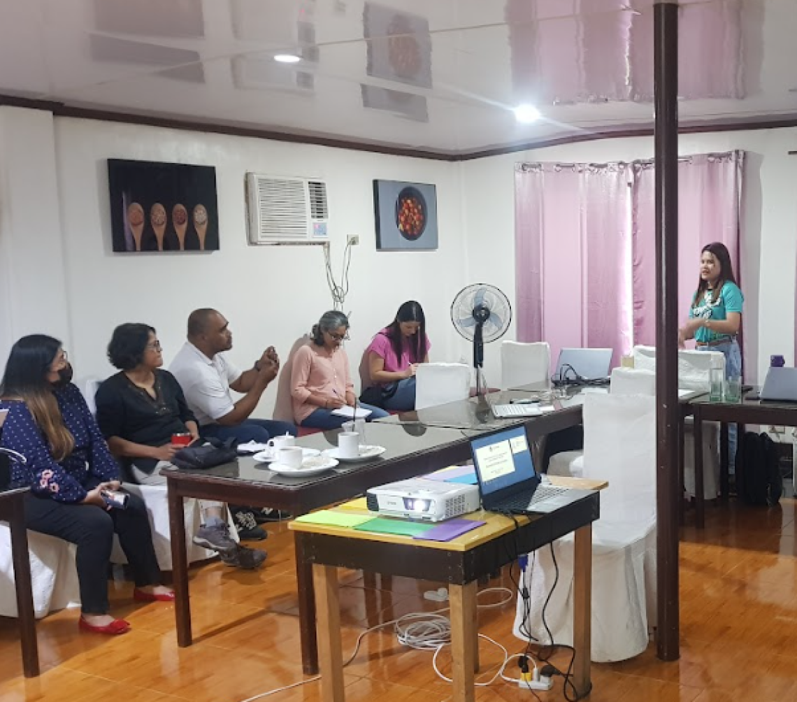 An indoor space with five people seated along a wall on the left side of the image. To the right, Michelle Laina stands and leads the conversation.