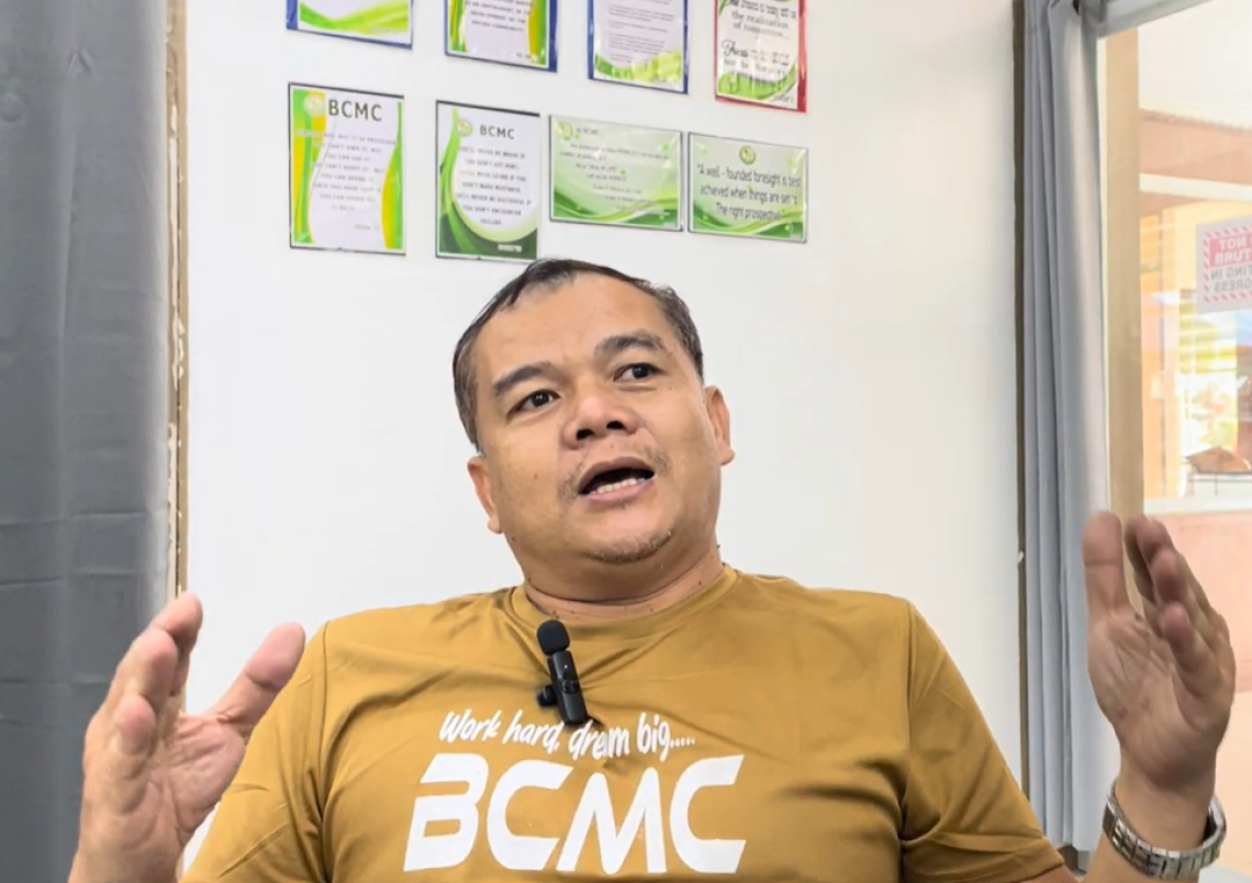 Image of Reynante Madelo, a man wearing a yellow shirt with short hair. Reynante has his hands in the air while leading a training session of copra farmers in the Philippines.