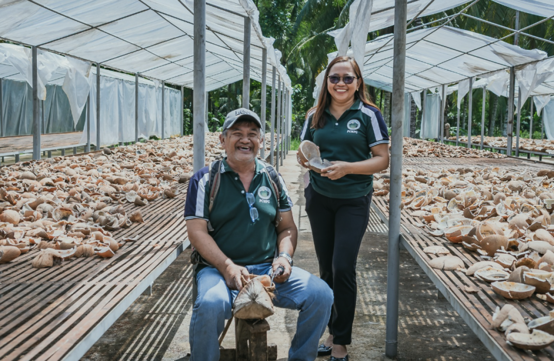 A man sits next to a woman standing. Both are in the middle of a coconut and copra drying facility.