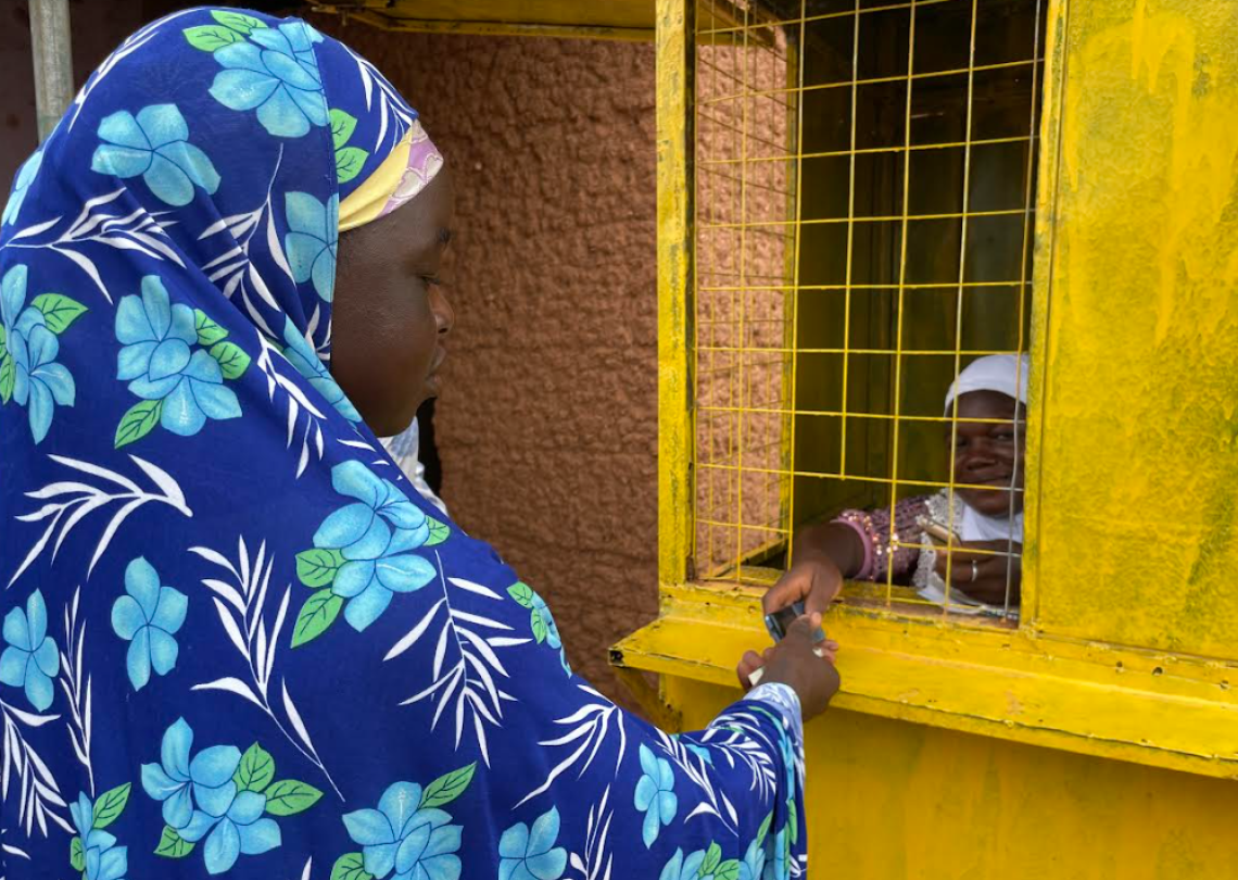 Ayisha, a mobile money agent in Ghana, assists her client, Fuseini, who is wearing a blue flowered head covering