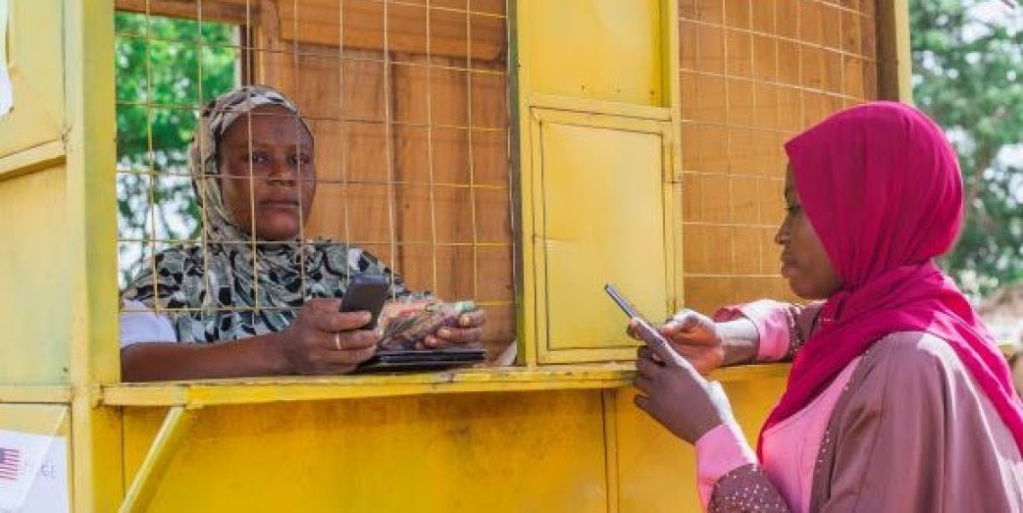 A Grameen Foundation DFS+ Agent assists a client in her kiosk. The client is wearing a pink headscarf and holding a phone.