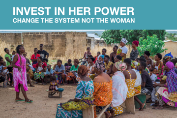 A woman stands in the center of a large group of seated people, teaching them about Grameen's power dynamics approach.  The audience is engaged with the speaker. All are outside and sitting under a tree.