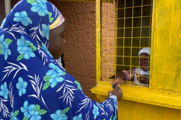 Ayisha, a mobile money agent in Ghana, assists her client, Fuseini, who is wearing a blue flowered head covering