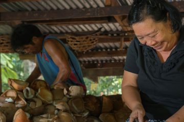 A woman cuts coconuts, a pile of shells in the background. A man is behind the pile, helping her.