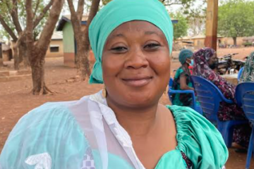 Sadia, a Grameen Agent in Ghana. She's sitting in a chair, wearing a turquoise headscarf and dress.