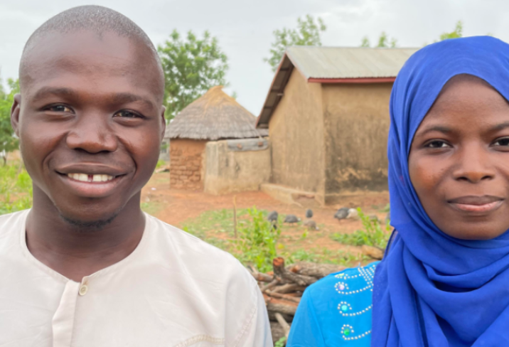 Yamba, from Northern Ghana, is wearing a white shirt. He stands next to his wife, Yahaya, who is wearing a blue headscarf and light blue dress.