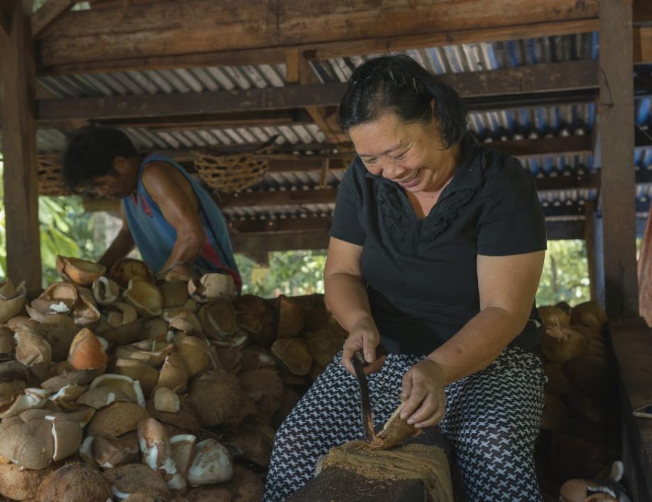 A woman smiles as she slices a coconut in half. A pile of coconuts, and a male farmer, are in the background.