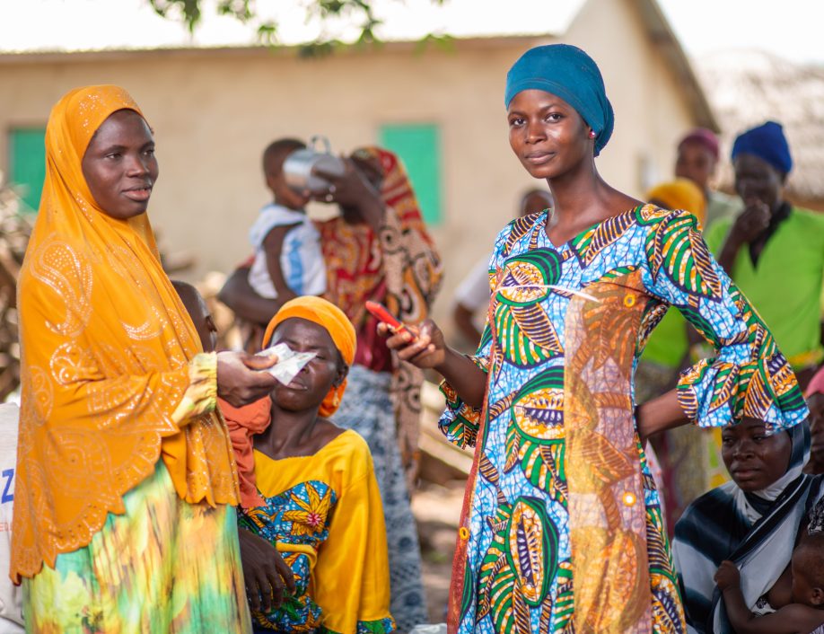 Two women stand outside with more people behind them. They are both dressed in bright colors and one is holding a phone. The other is completing a mobile money transfer. Grameen Foundation works to end poverty by bringing financial services to people living in rural locations.