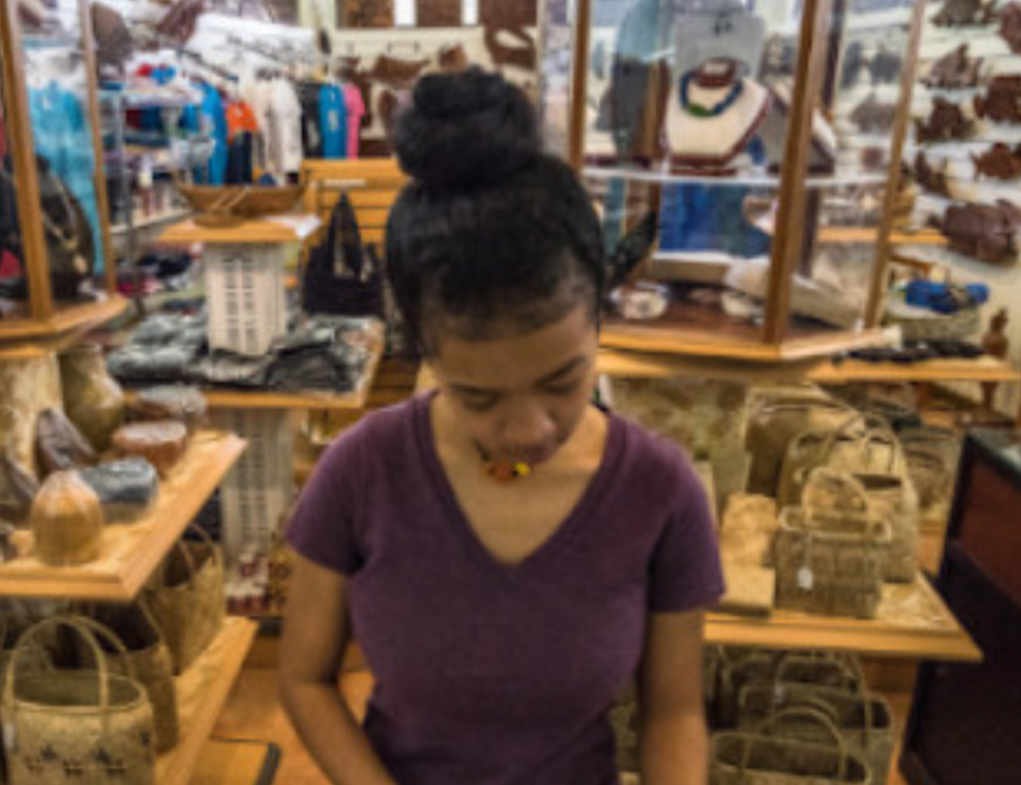 An image of a woman standing in a shop. She has a purple shirt on, dark brown hair pulled into a bun on the top of her head, and is looking down at a table