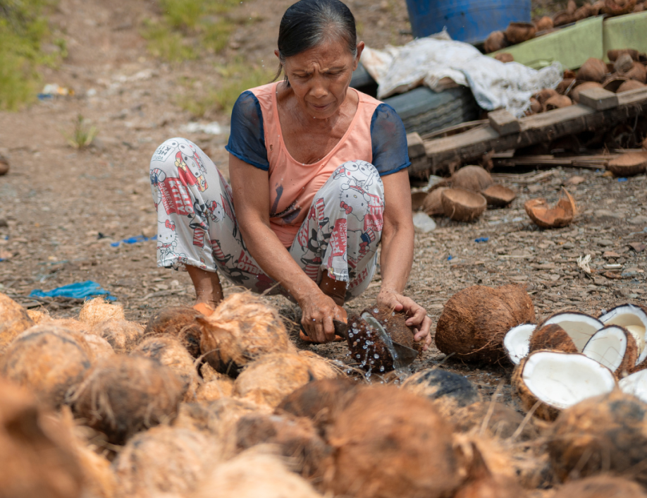 A woman is crouched down near the ground and cutting open a coconut. Water is splashing out of the coconut and there are is a large pile of coconuts in front of her.