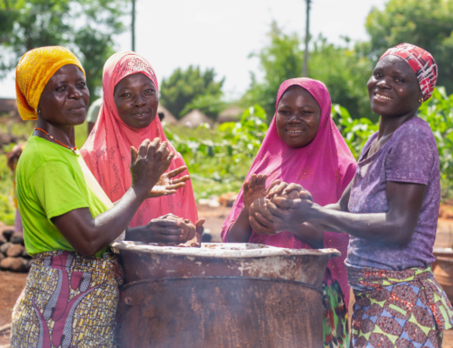 A group of women stand at an outdoor table -all are smiling at the camera and working together.