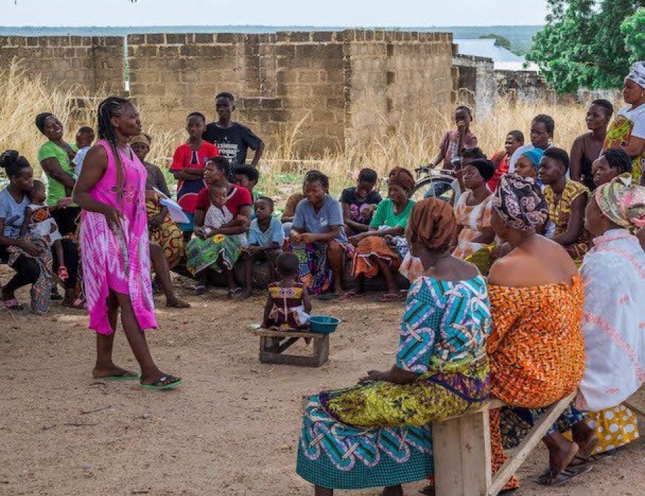 Patience stands in front of a large group of people, seated outside. Grameen Foundation works with women like Patience to become leaders in their communities.