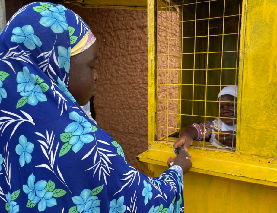 Ayisha, a mobile money agent in Ghana, assists her client, Fuseini, who is wearing a blue flowered head covering